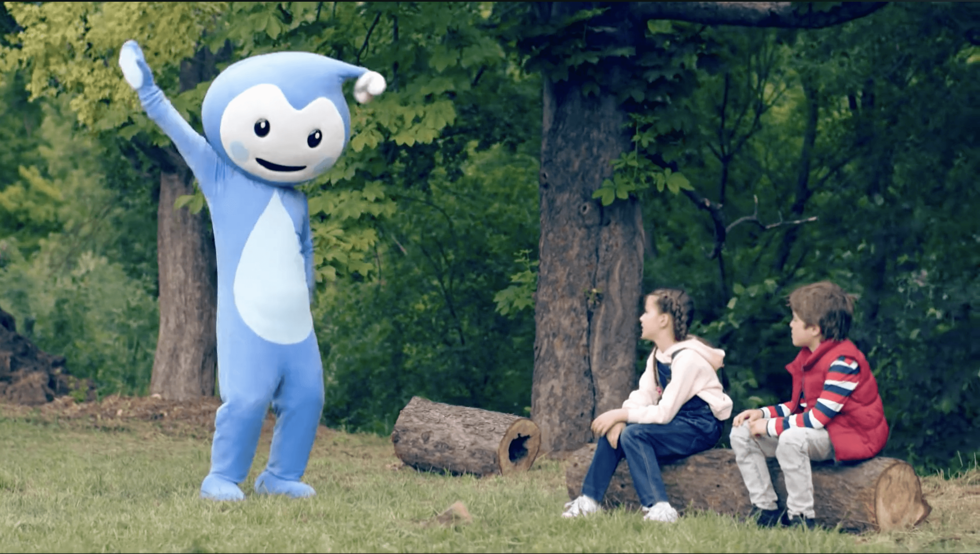 Cseppbarát kékl kabalafigurája táncol az erdő szélén a gyerekek figyelik egy tuskón ülve.