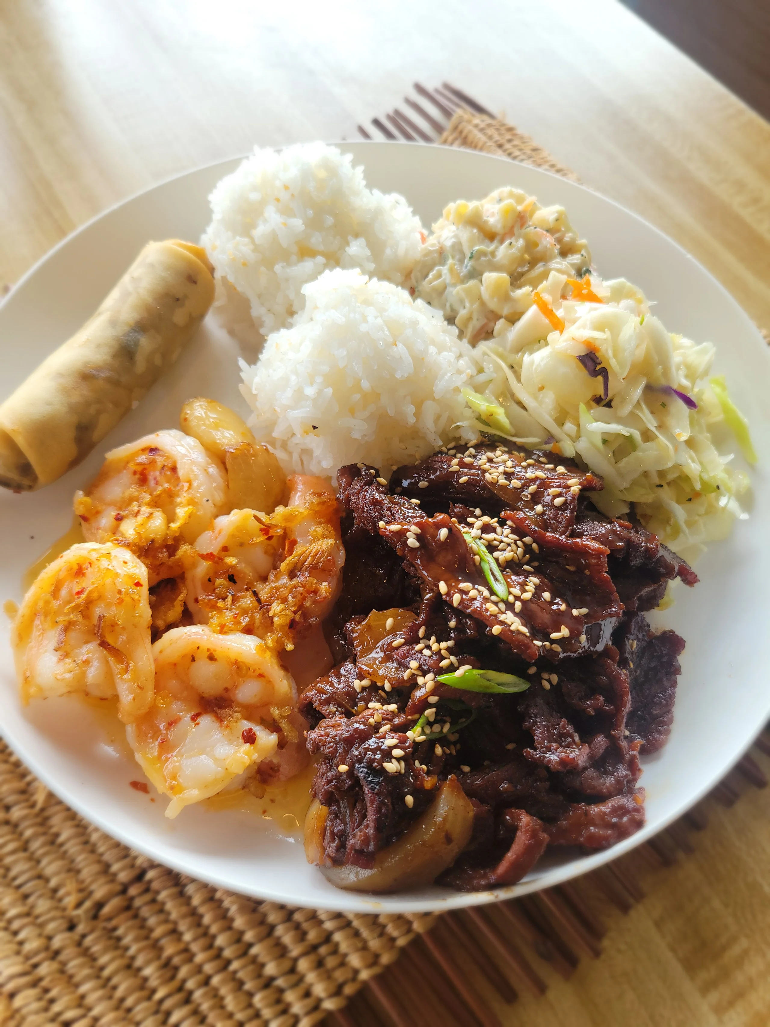A large white plate featuring thin-sliced teriyaki beef and four large garlic shrimp, served with a spring roll, white rice, macaroni salad, and slaw.