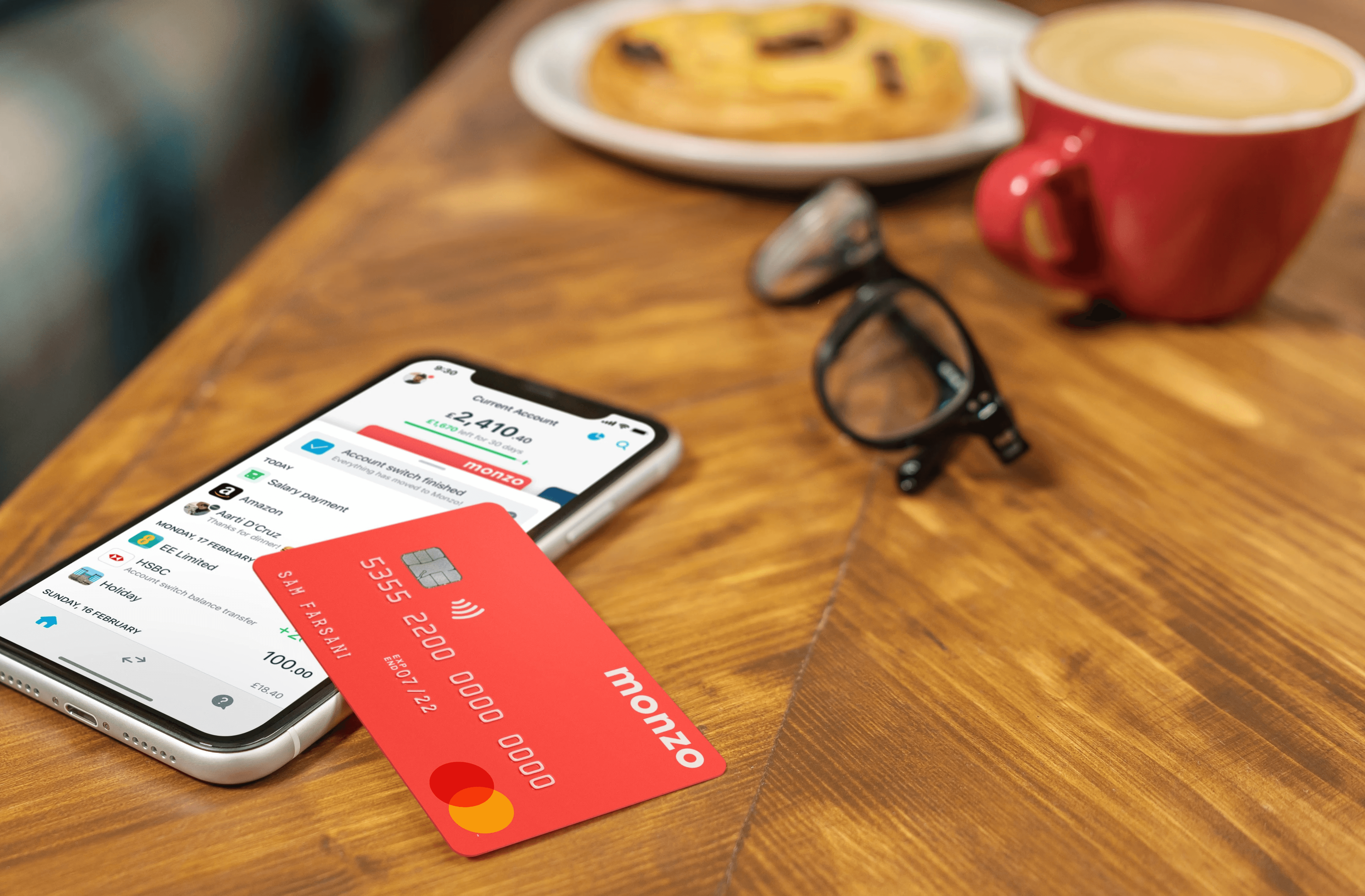 Image of a mobile phone on a table with a Monzo account showing on the screen, and a Monzo bank card on top of the phone at an angle. The setting is in a café, with a coffee, cookie and glasses on the table near the phone and bank card.