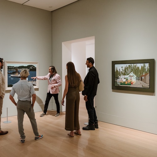 一群人正在欣賞藝術畫廊中的畫作。一位穿著粉紅色外套的人朝著一幅海岸場景的畫作比劃。