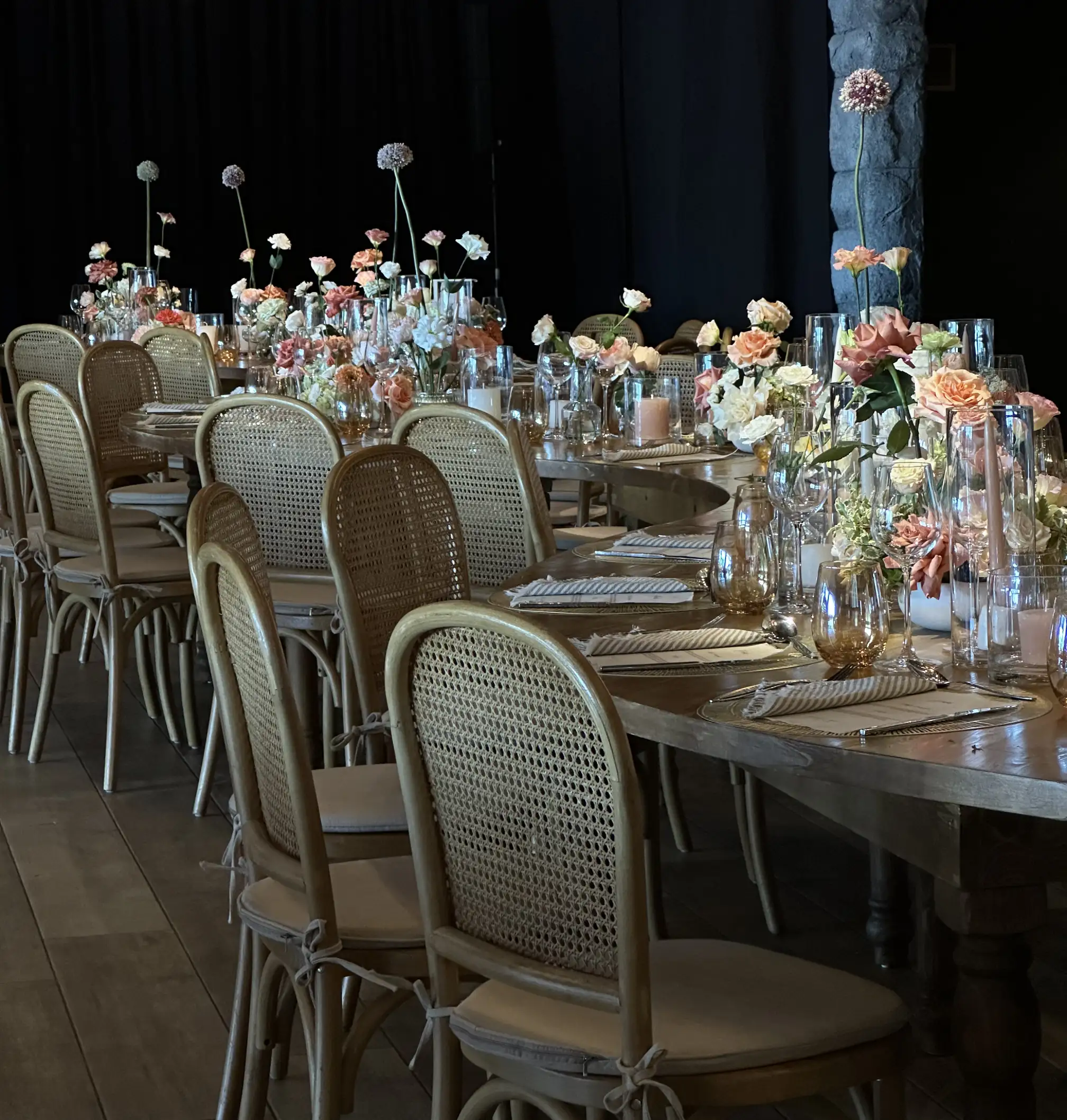 Mesa larga decorada con flores en tonos pastel, velas y cristalería fina, rodeada de sillas de ratán en un ambiente elegante y cálido.