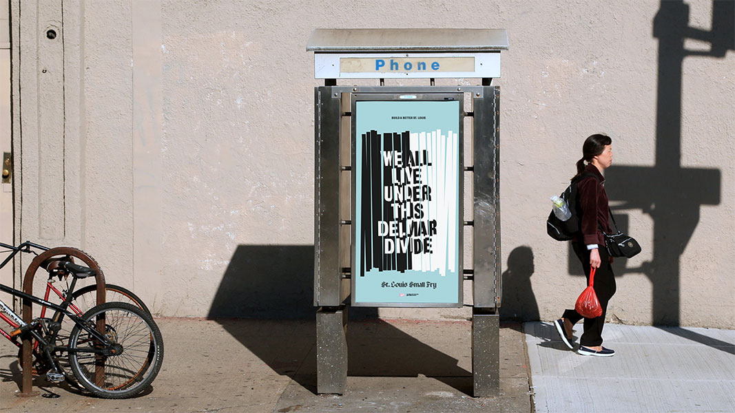 A woman walks past a phone booth on a sidewalk. She carries a backpack and holds a red bag. Two bicycles are parked nearby against a light-colored wall. The phone booth displays a poster with text reading “WE ALL LIVE UNDER THIS DELMAR DIVIDE.” The poster includes text mentioning “St. Louis Small Fry” and “ST. LOUIS SMALL FRY.”