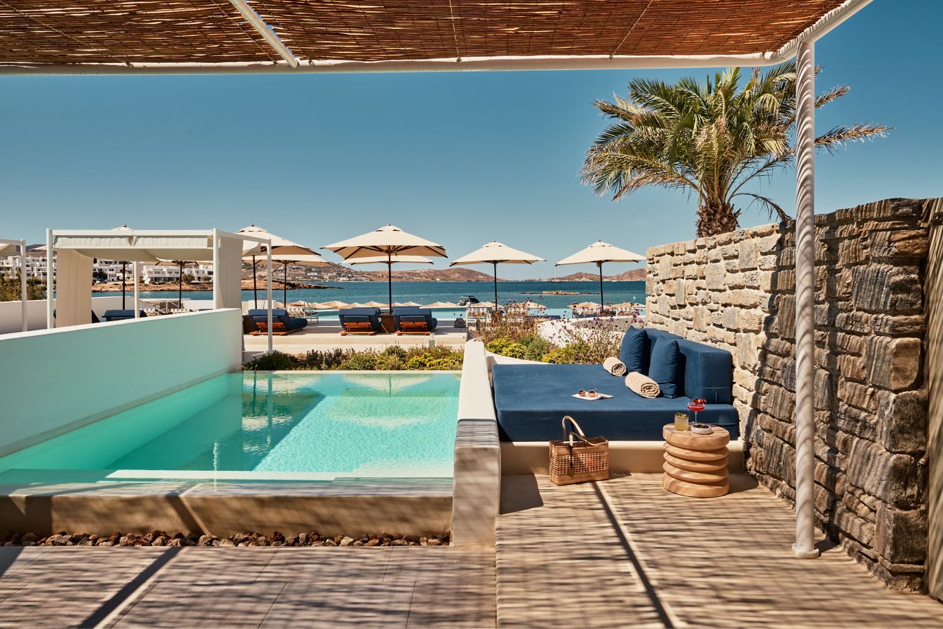 Luxurious hotel terrace with a private pool, a large blue daybed, and a view of the sea with umbrellas in the background.