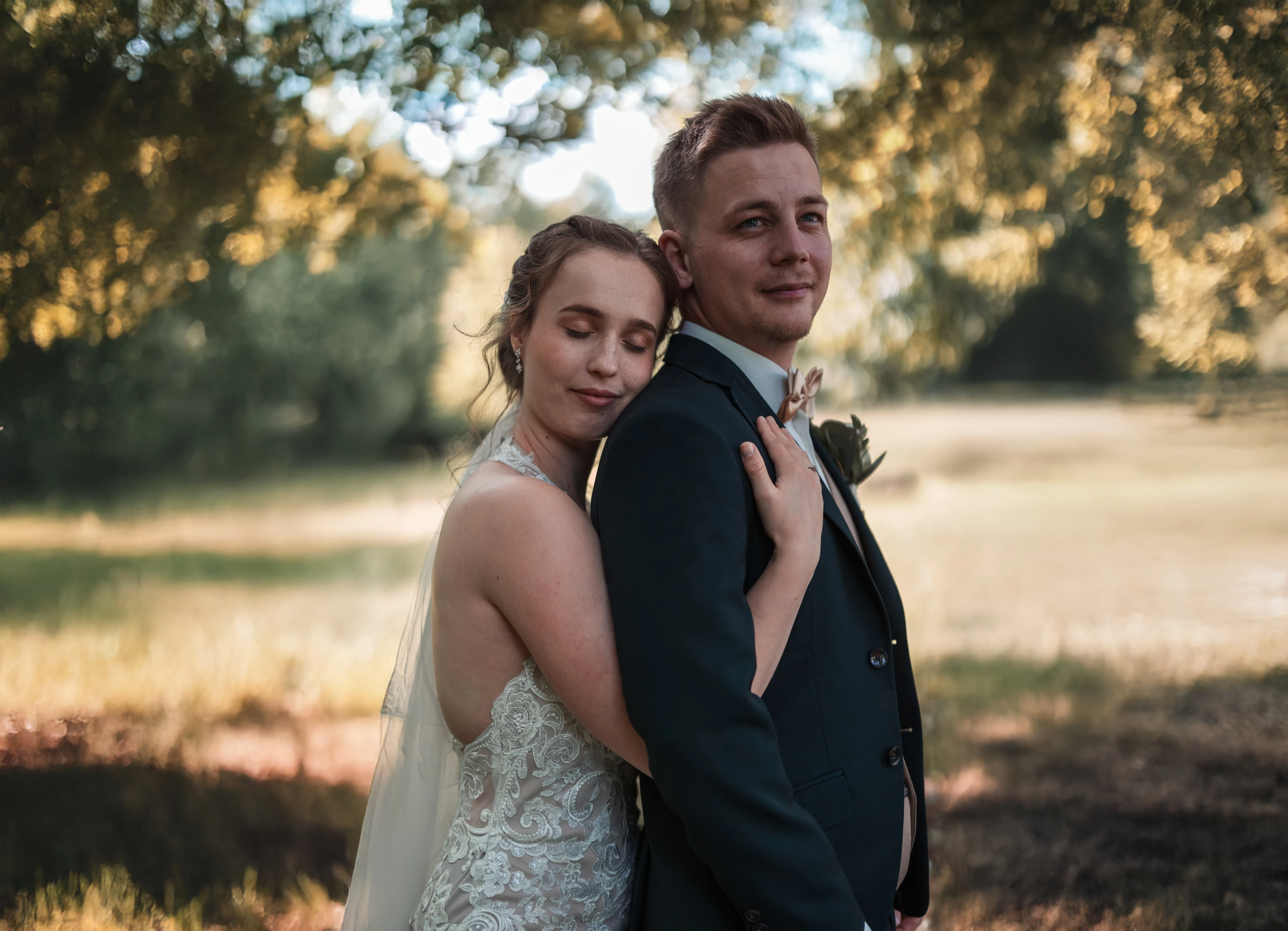 Romantikus esküvői portré: menyasszony csipkés ruhában és fátyollal hátulról öleli vőlegényét fekete öltönyben parkban, meleg aranyóra megvilágításban Nagyatádon
