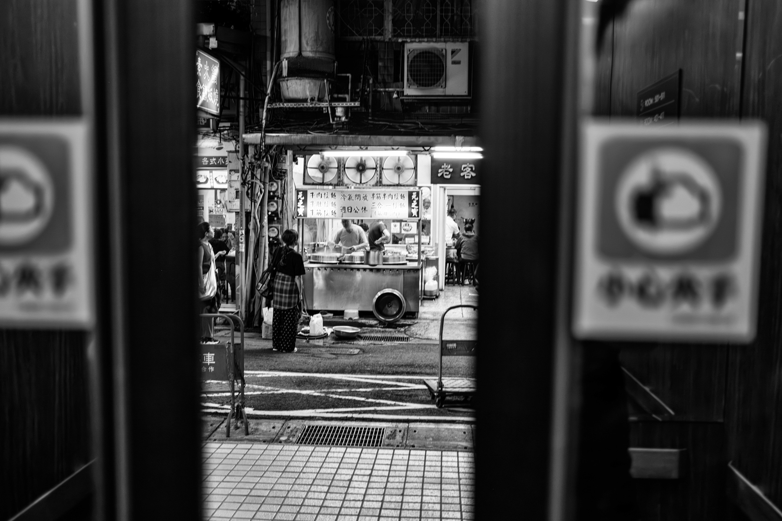 Black and white photograph framed through two doorways showing a street food stall with customers waiting to order.