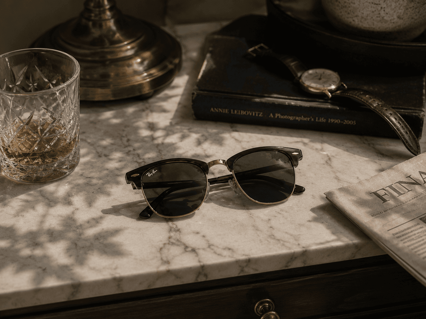 High-angle view of classic tortoiseshell clubmaster sunglasses with dark lenses resting on a marble-top table. The vintage-style setting features an Annie Leibovitz book with a leather-strap watch on top, a crystal glass with an amber liquid, a corner of a newspaper, and soft leaf shadows cast across the surface.