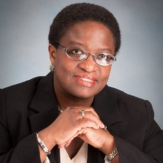 A portrait of a black woman with close cropped hair, rimless glasses, and silver earrings, wearing a black suit coat over a pink blouse, her hands are clasped in front of her