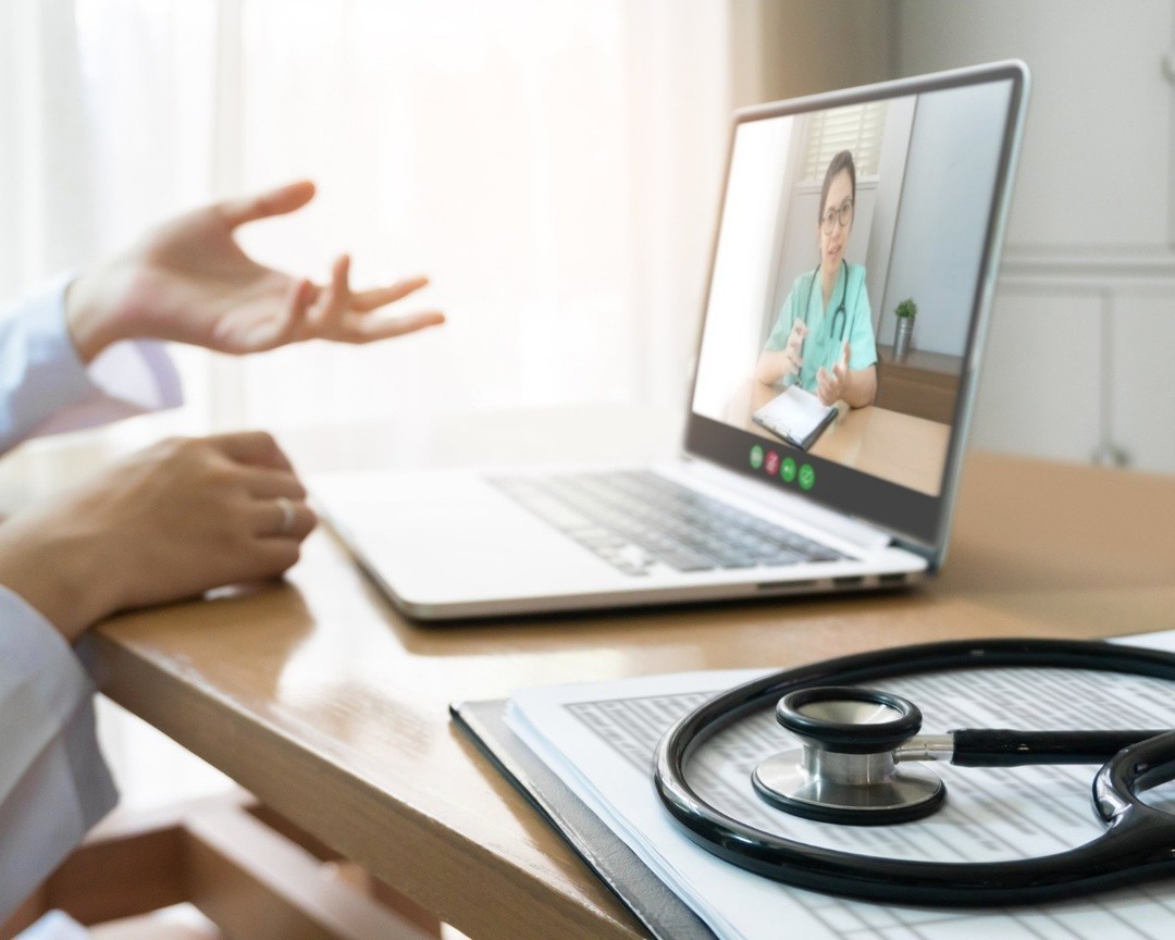 Patient in video call with doctor on laptop during telehealth visit, with stethoscope and medical chart on desk.