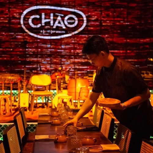 Seorang individu menyusun meja dalam sebuah restoran yang redup dengan perkataan "CHÁO" terang di dinding bata latar belakang.