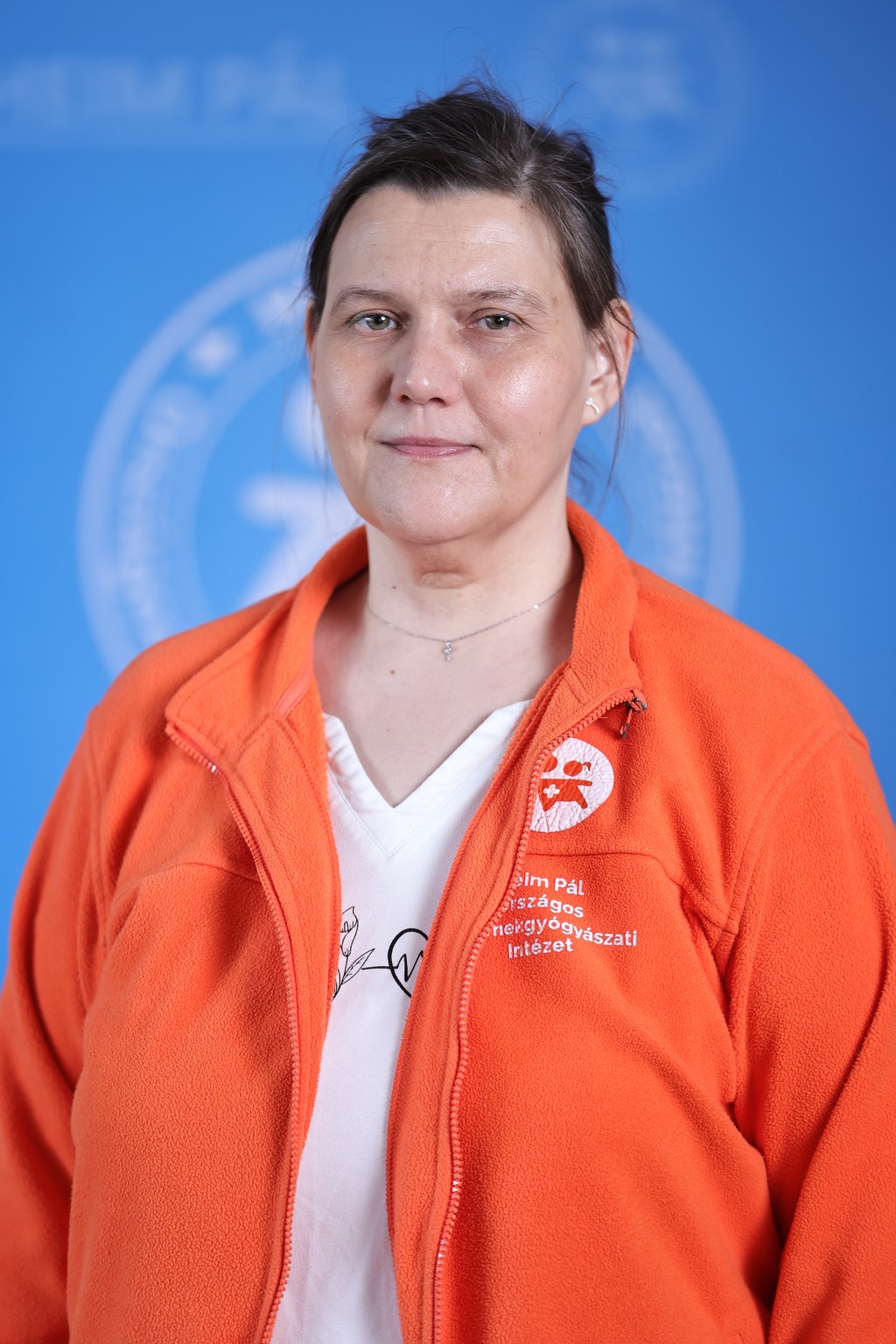 Világos hajú nő narancssárga heim pál logós pulóverben kék háttér előtt.