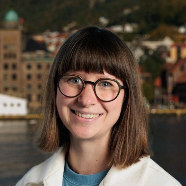 Smiling young man with curly hair, wearing a dark hoodie, against a softly lit background.