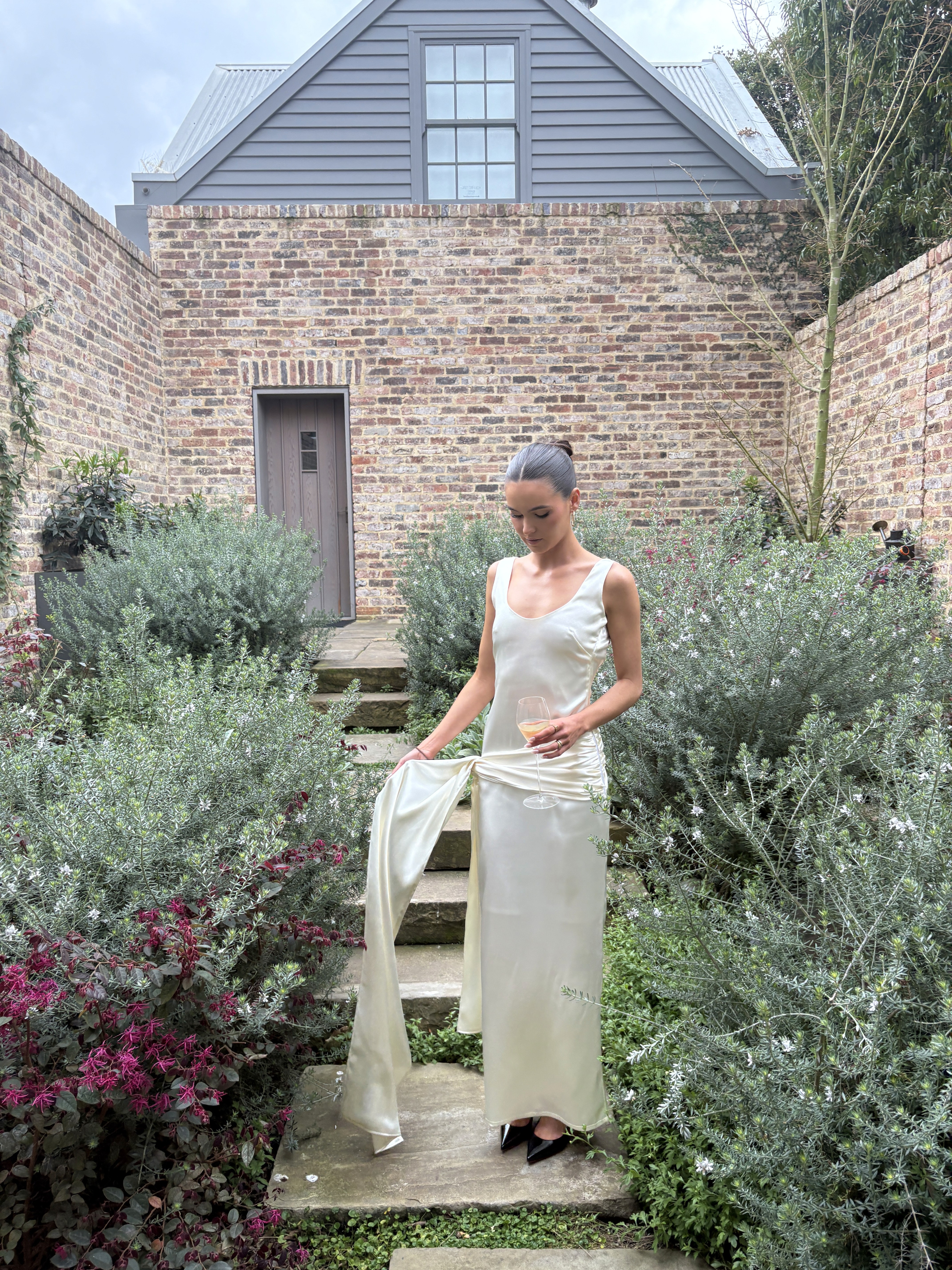 Floor-length ivory satin slip dress with scoop neckline, sleeveless cut, and asymmetrical draped sash detail at the hip