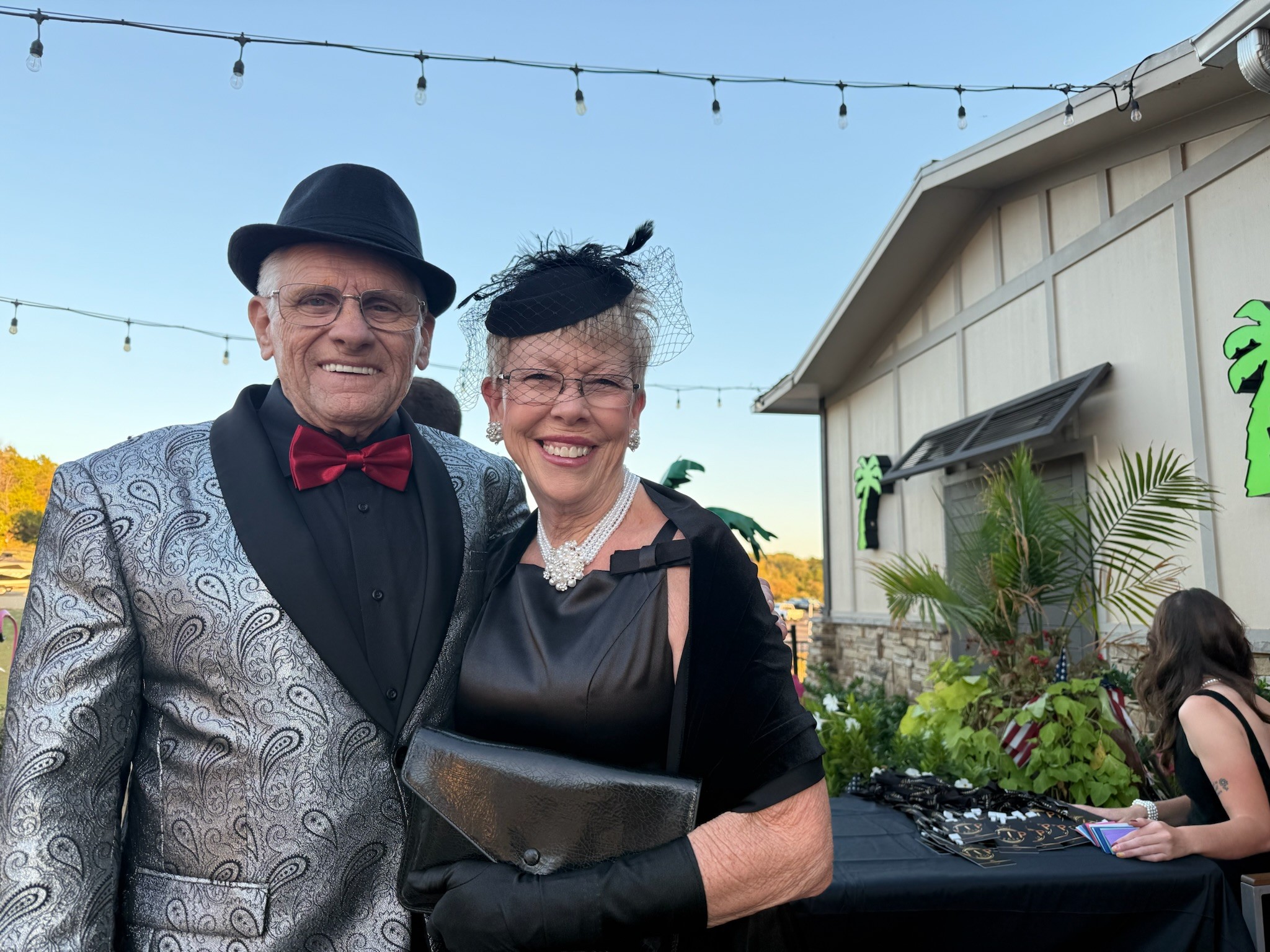 A smiling elderly couple dressed in elegant evening attire, with the man in a silver paisley jacket and black hat, and the woman in a black dress and fascinator, pose at an outdoor event under string lights next to a booth with decorations.