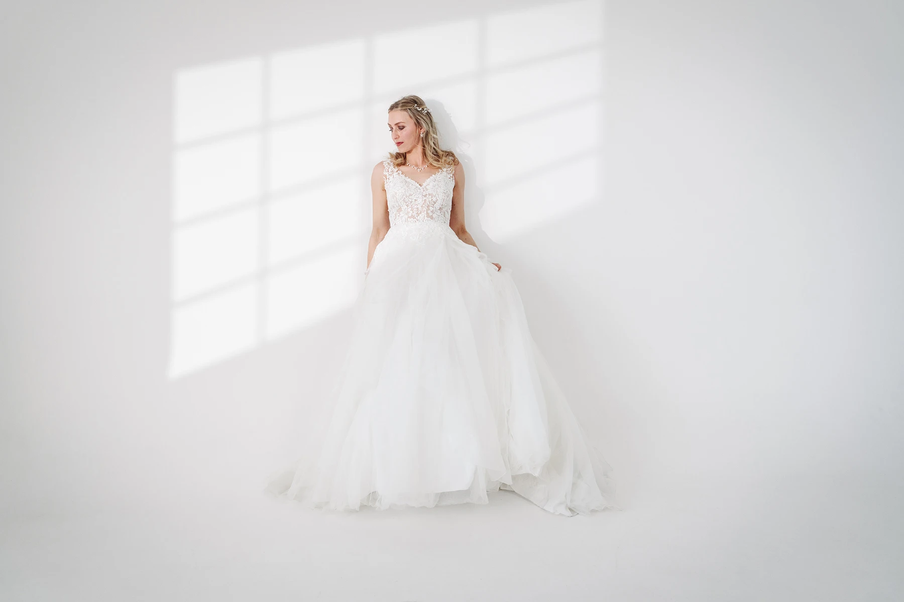Braut in weißem Spitzen-Hochzeitskleid steht vor einem Fenster im Fotostudio Erfurt, mit sanftem Licht und Schatten.