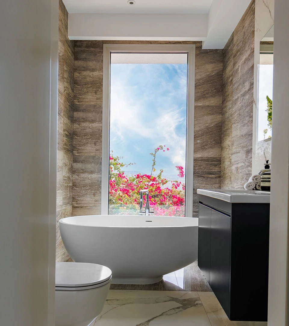 Seafront Residence Limassol bathroom with freestanding tub, stone-textured walls, and picture window framing garden blooms