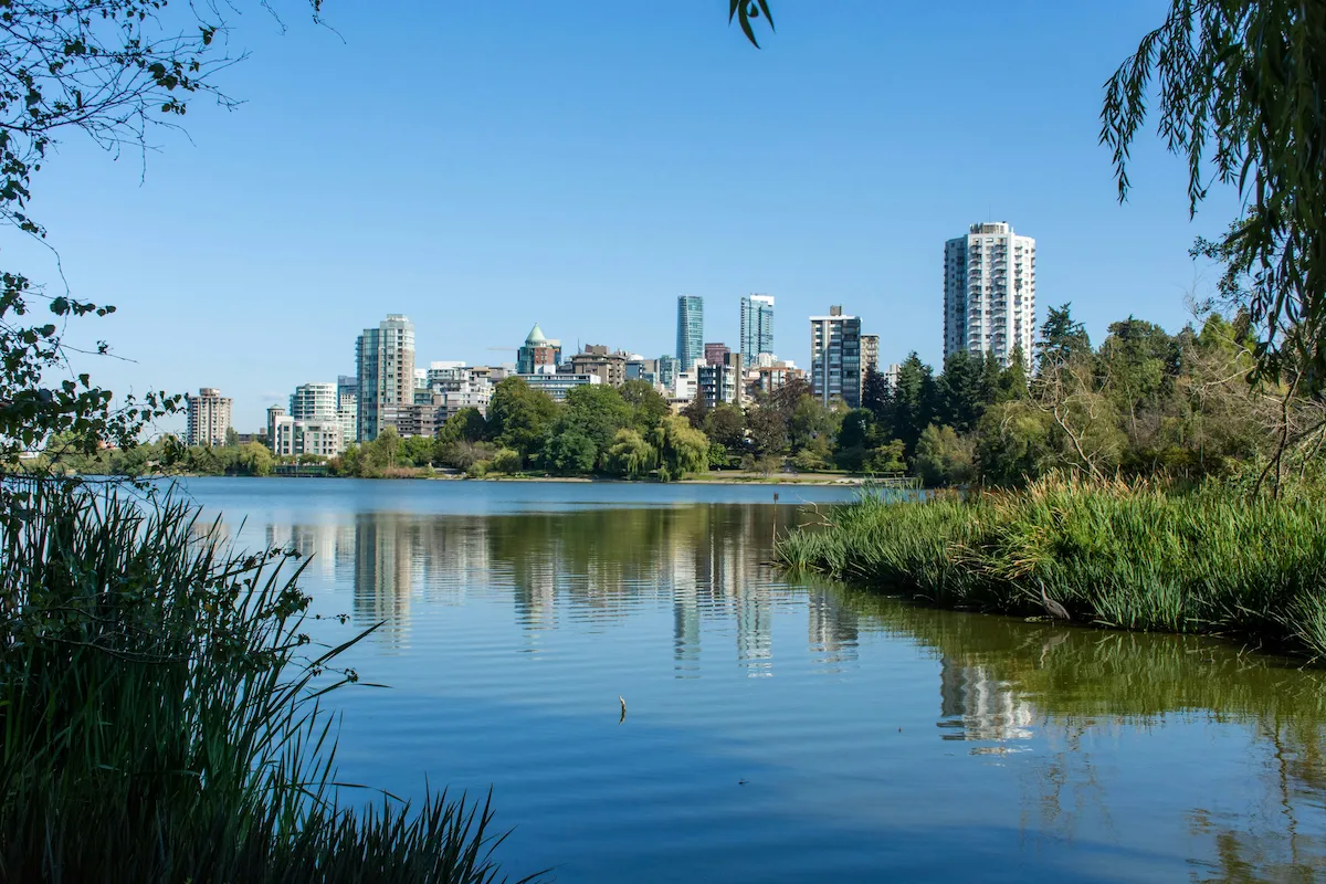 Cityscape of Surrey, British Columbia, viewed across a calm lake with surrounding trees and high-rise buildings in the background.