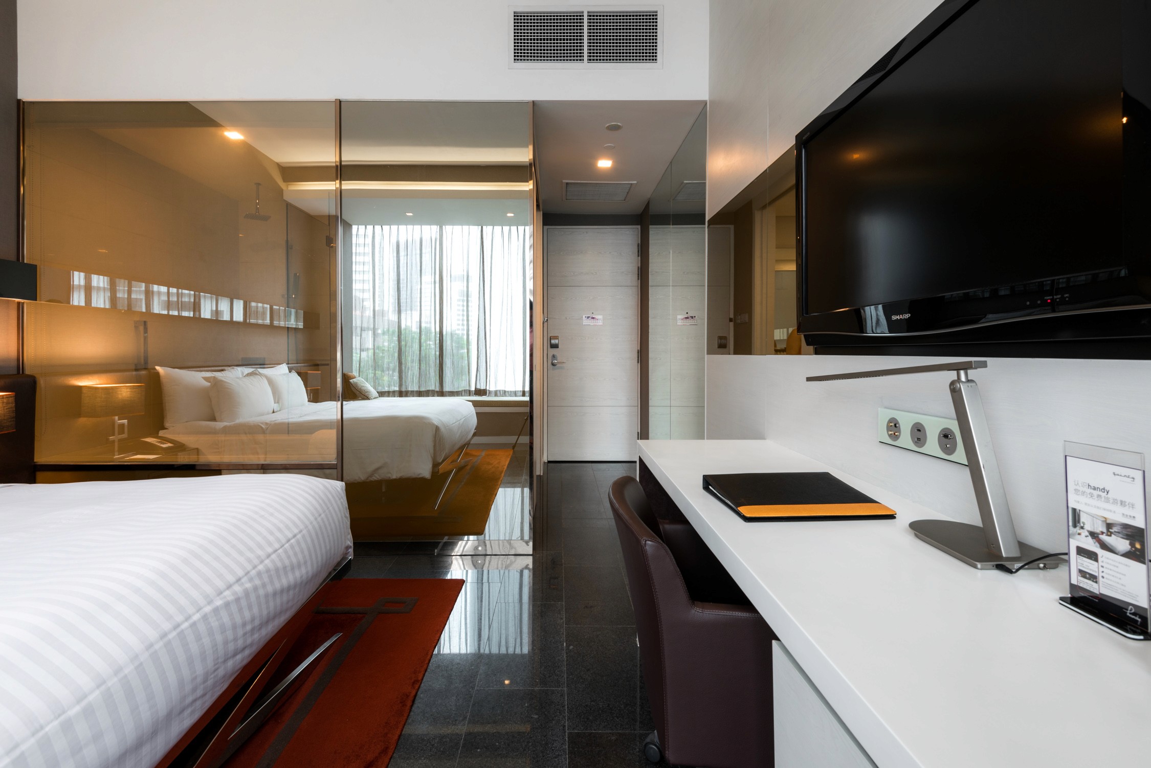 A modern hotel suite with two beds and a white desk area featuring a television and a laptop in the foreground.
