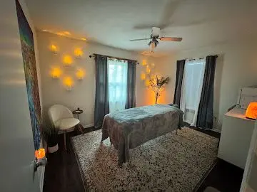 A serene massage room in Bradenton, FL, features a neatly made massage table adorned with a gray blanket, ambient wall lighting casting a warm glow, sheer and dark drapes on windows, a cozy chair, and a decorative salt lamp, enhancing the tranquil atmosphere.