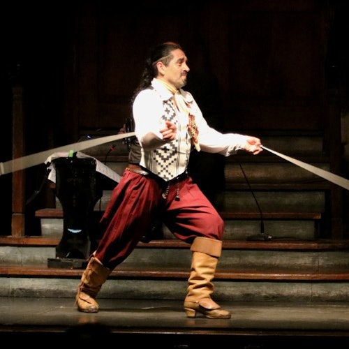 Un hombre con atuendo tradicional actúa en el escenario, extendiendo una cinta con ambas manos. En el fondo se ven unas escaleras y un soporte decorativo.