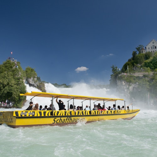 A yellow boat filled with people cruises near a waterfall, surrounded by green trees and a clear blue sky.