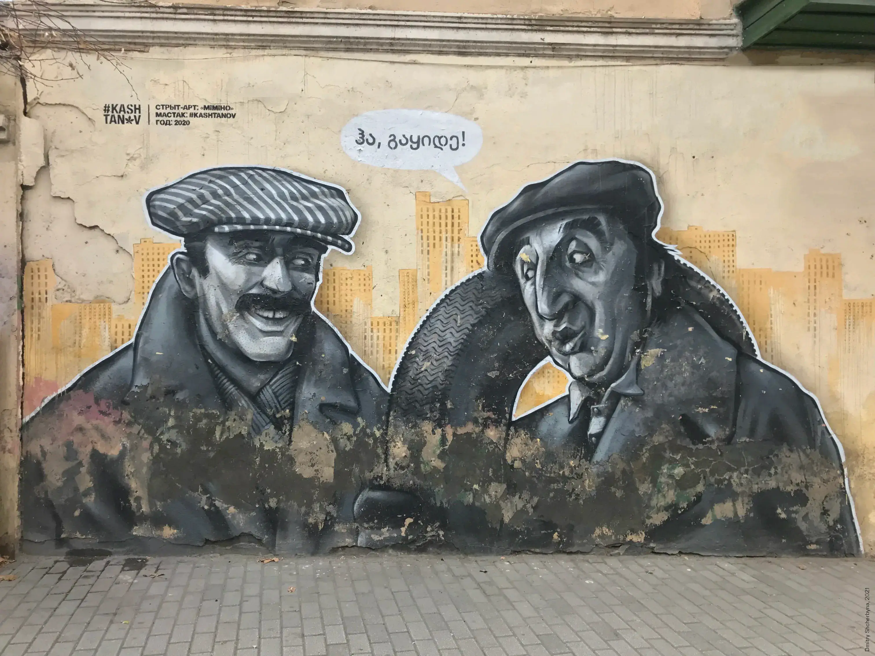 A grayscale mural of two men in coats and hats smiling and leaning towards each other, with a golden city skyline behind them. They are characters from the film "Mimino".