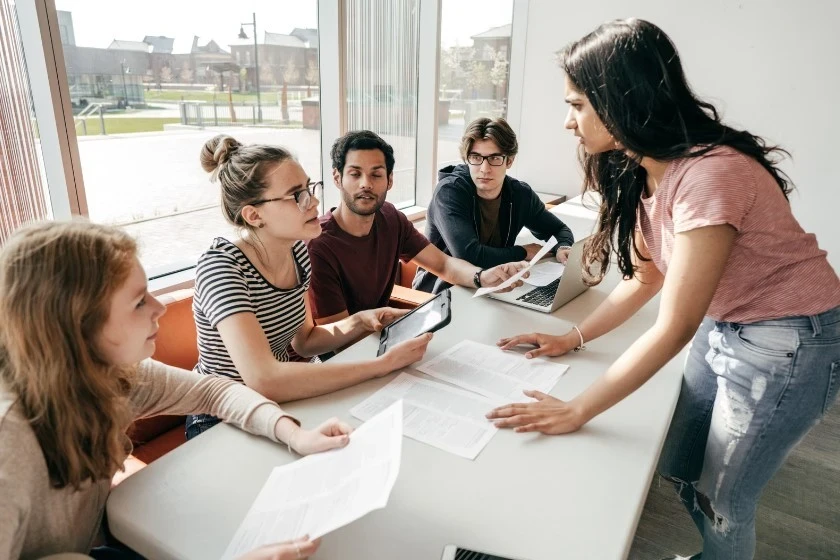 Teens engaged in a collaborative learning session on anthropology, with one student presenting ideas while others follow with laptops and notes.