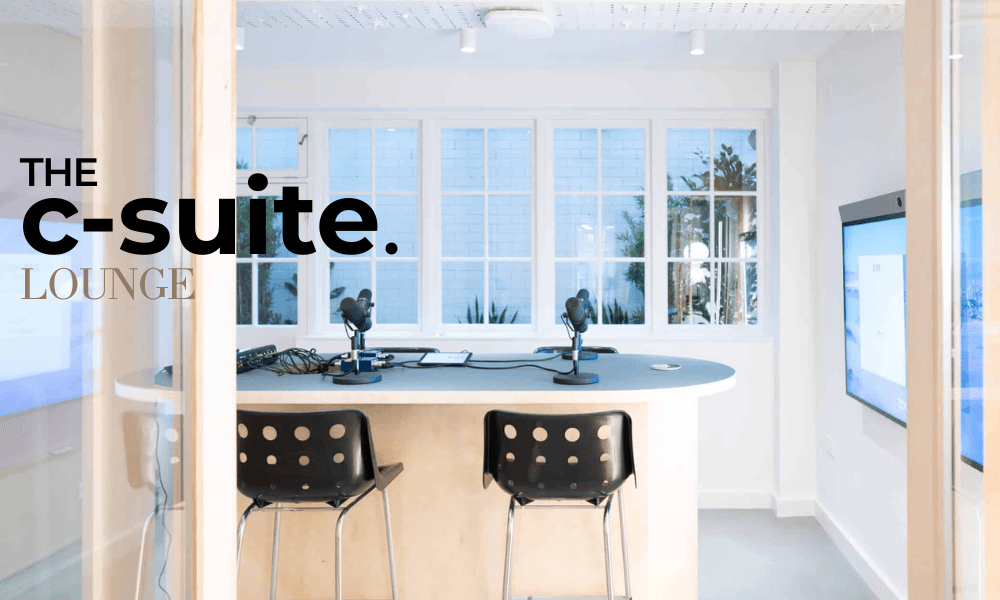 C-Suite-Invitationer and a woman in a light sweater sit at a desk with a laptop, discussing something. The room is dimly lit, creating a focused atmosphere.