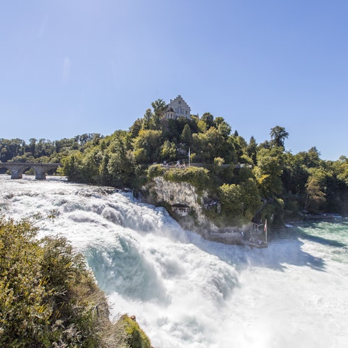 A large waterfall surrounded by lush greenery and trees with a castle-like building on a hilltop under a clear blue sky.