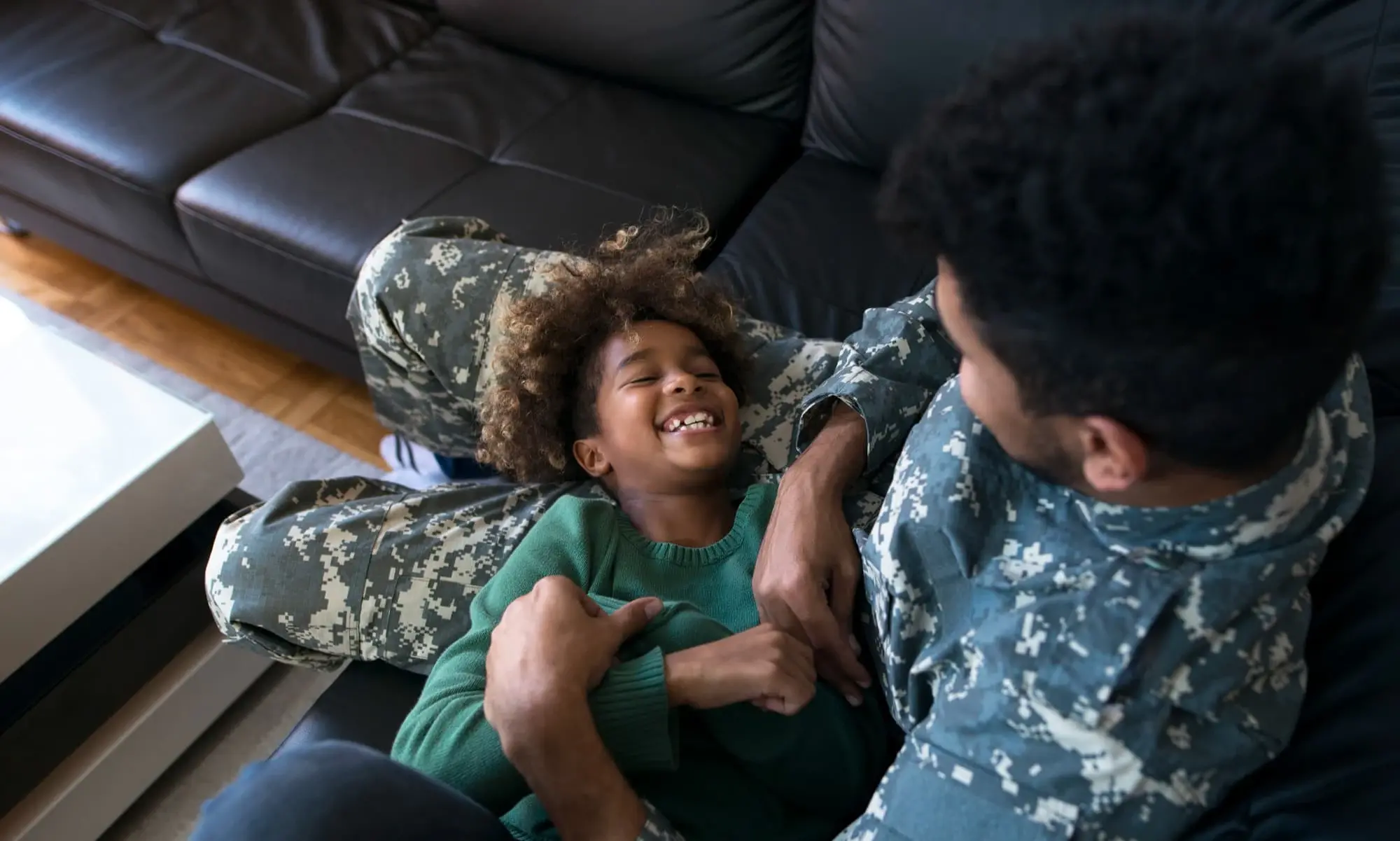 A military parent in uniform sits on a couch laughing and playing with their young child. The joyful moment represents family connection, gratitude, and the homeownership benefits made possible through VA loans from Chris Lewis Home Loans.