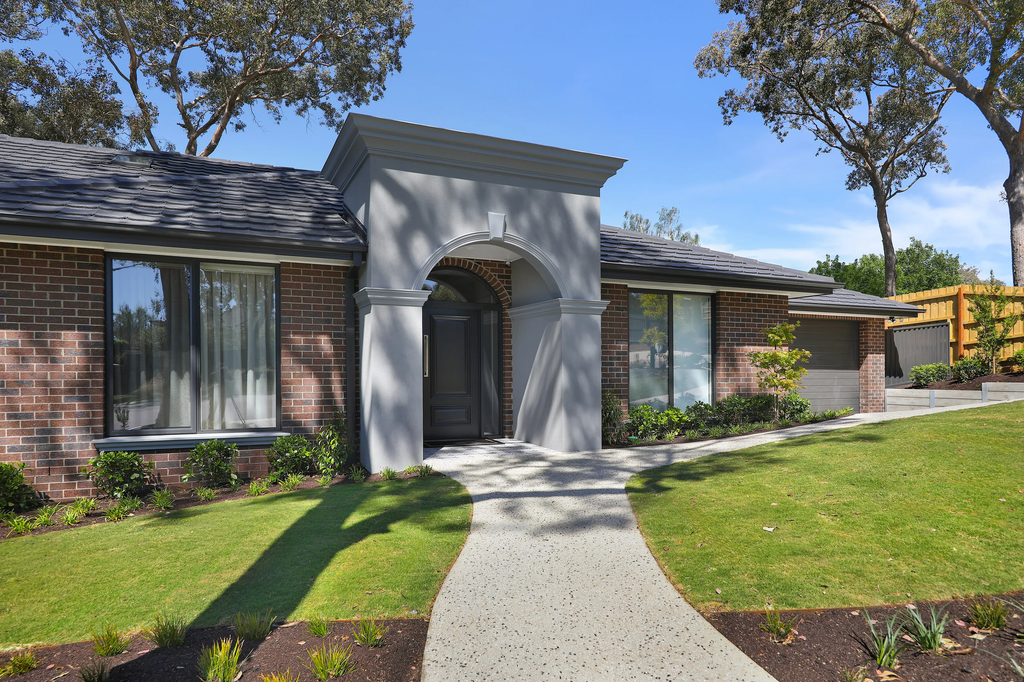 Front of the Diamond Creek home with a rendered arched entry portico, blend-brick walls, dark grey tiled roof, charcoal-framed windows, an aggregate path and a brick mailbox numbered 8