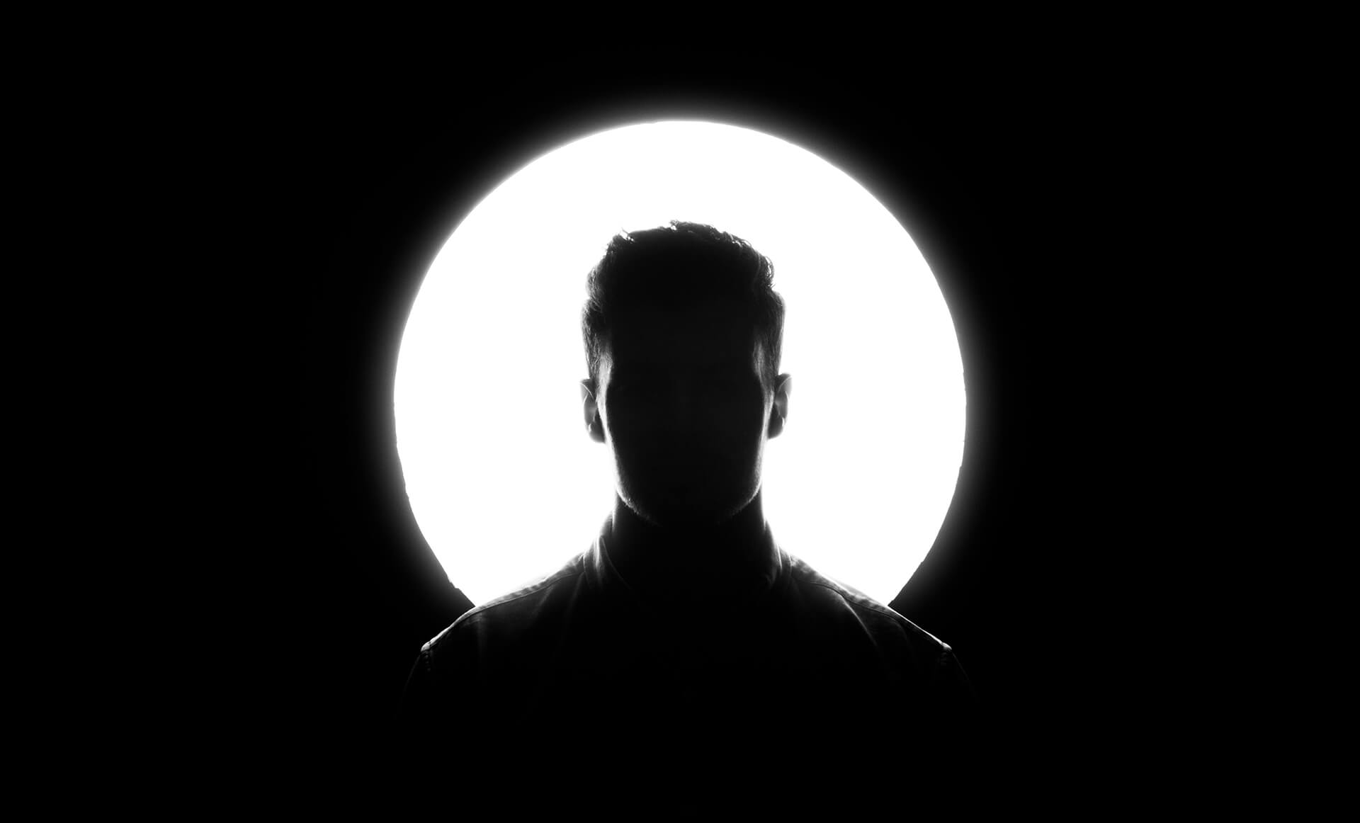 A silhouette of a man is centered against a bright, circular white light, creating a dramatic halo-like effect. The background is entirely black, emphasizing the contrast between light and shadow.