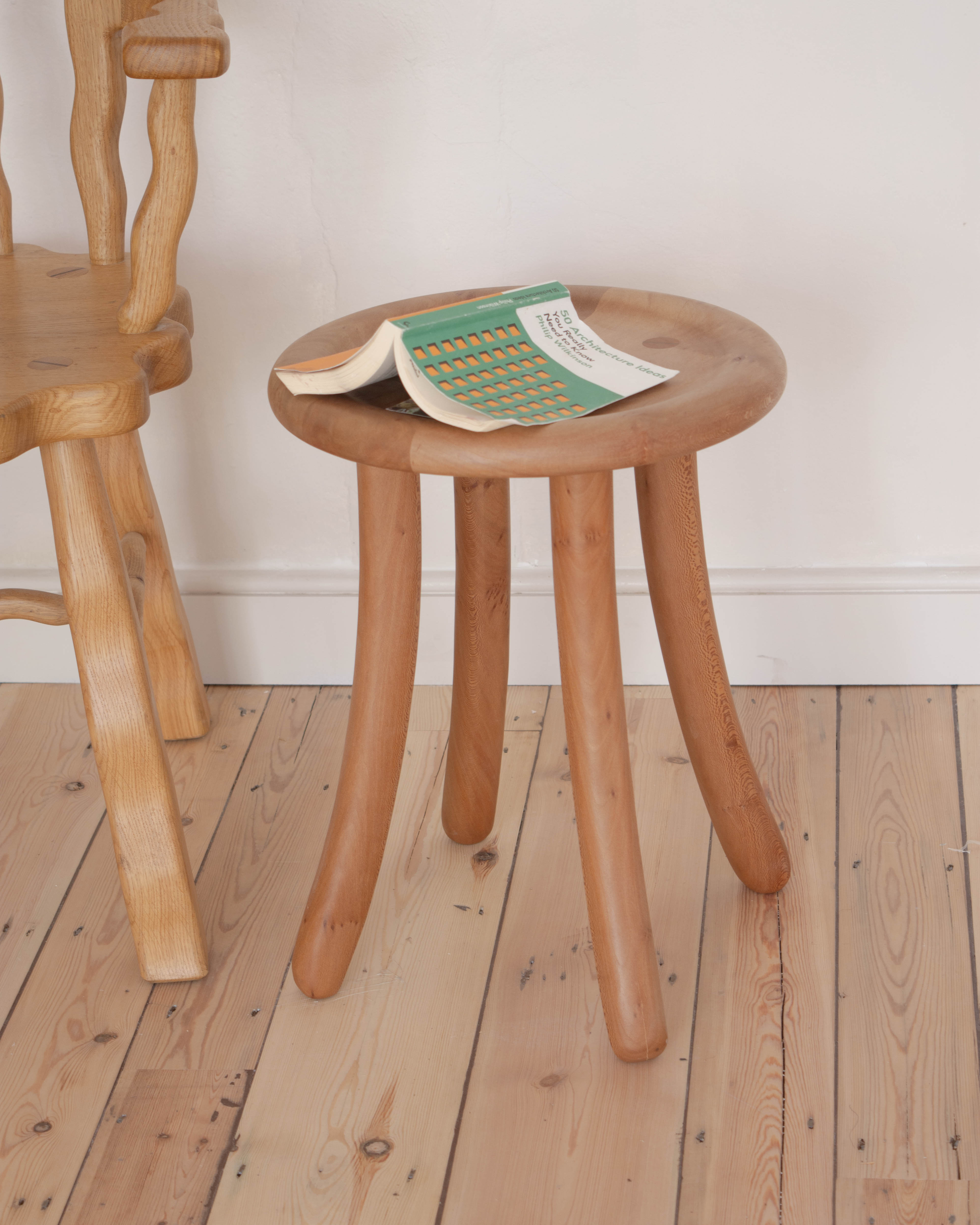 Wooden curved Luneta stool with a book on a wooden floor