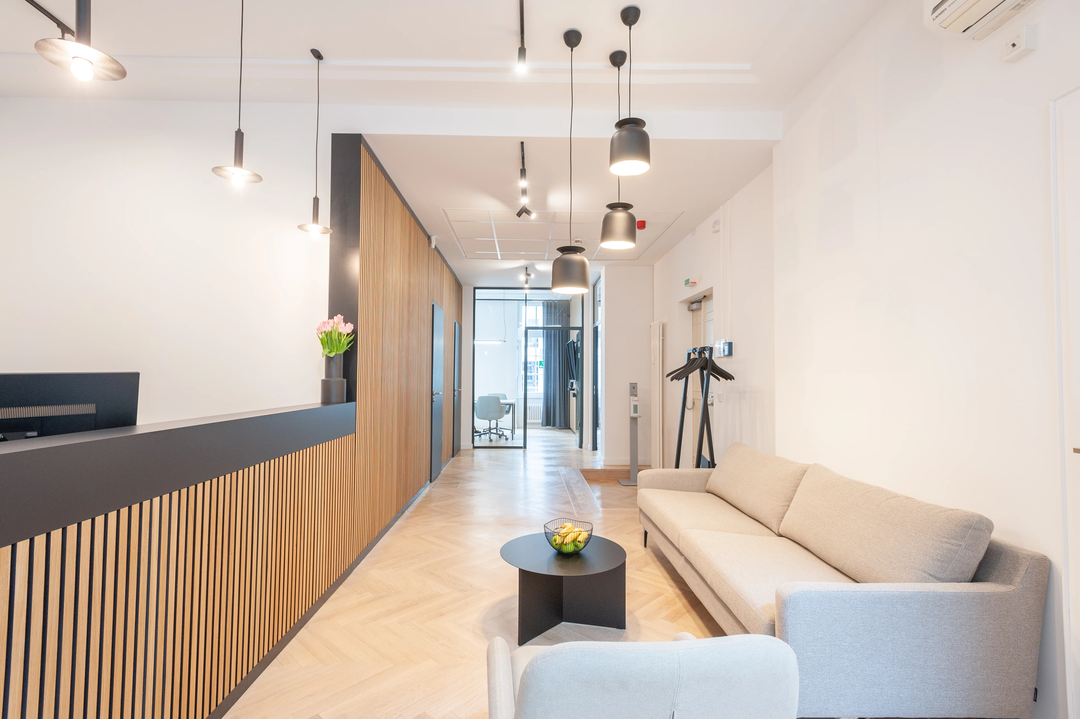 Reception corridor at AI START-UP BERLIN with a black counter on a wooden slat base, black pendant lights, a beige sofa with black coat stand and herringbone parquet flooring.