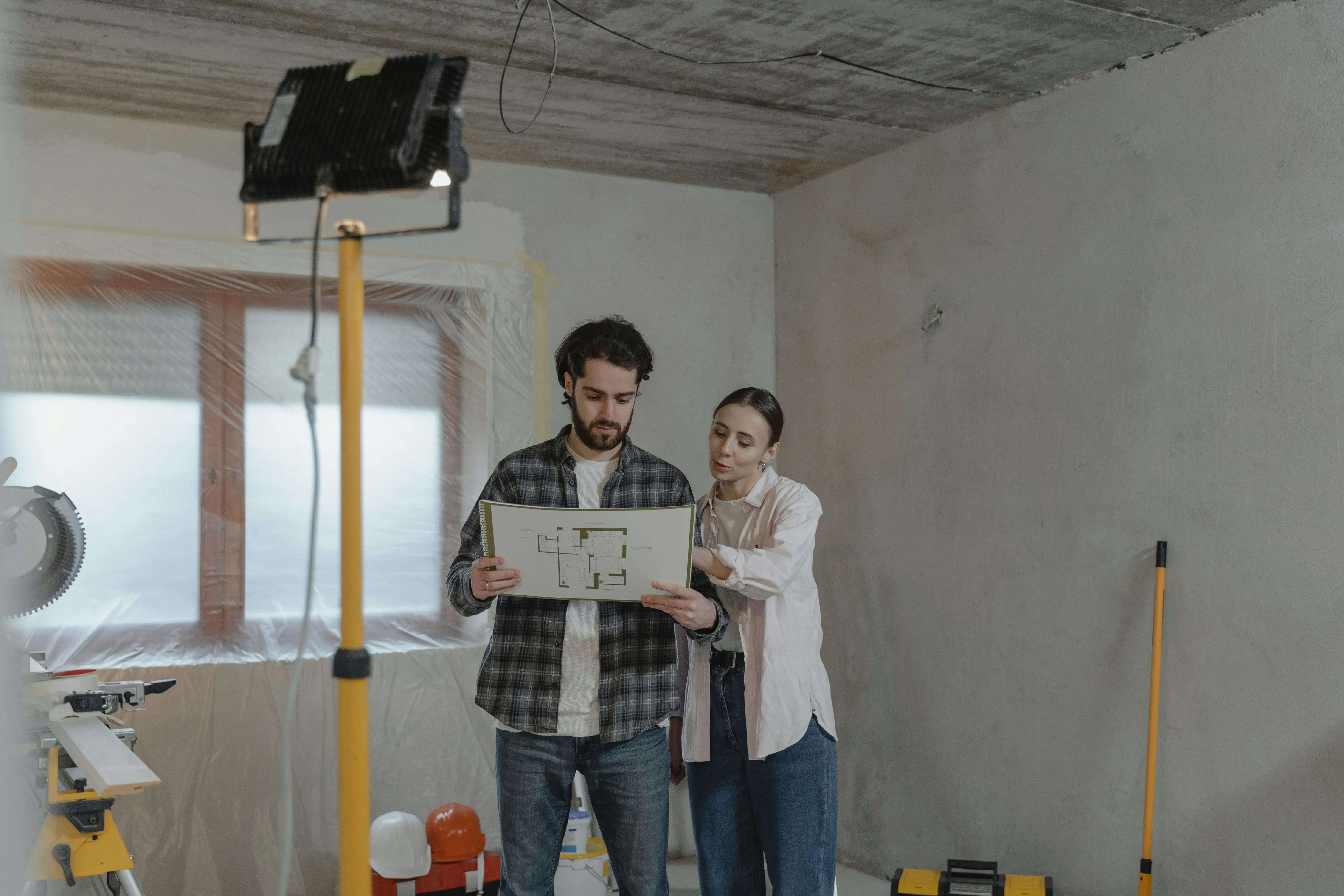 A man and woman in a partially renovated room study blueprints. They appear focused, surrounded by construction tools, wrapped windows, and exposed walls.
