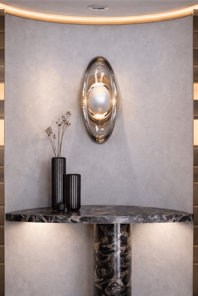 Luxury bronze and glass wall sconce mounted on a textured plaster wall above a marble console table with decorative vases in a modern minimalist interior.