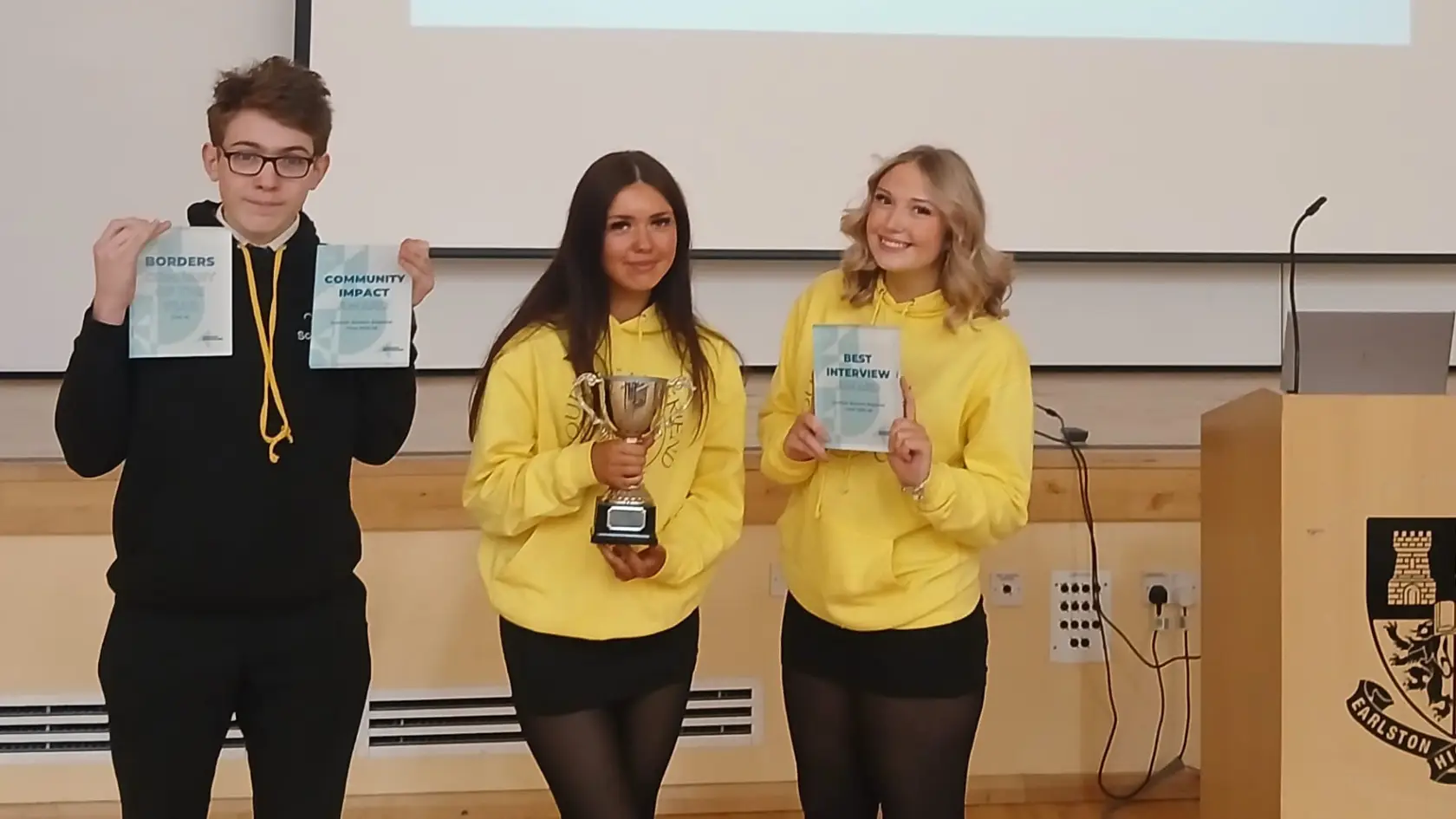 Three smiling people stand in a row. The left and right individuals hold certificates, while the person in the middle holds a trophy. They wear yellow tops.