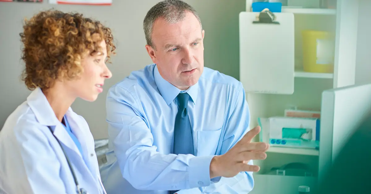 Male doctor explaining information to a colleague while pointing at a computer screen in a clinical office