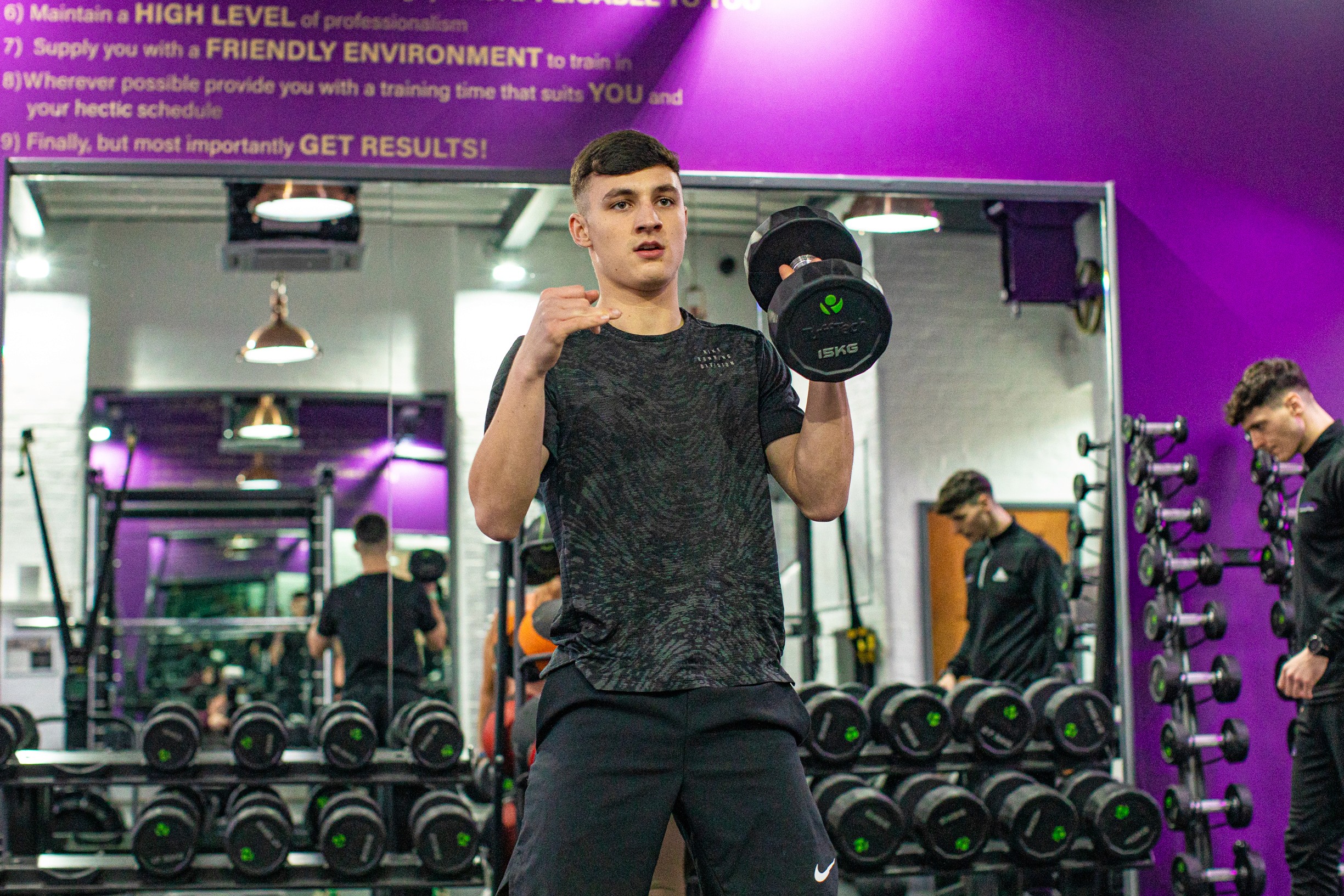 A person lifting a dumbbell in a gym, surrounded by weights and fitness equipment, with a purple wall.