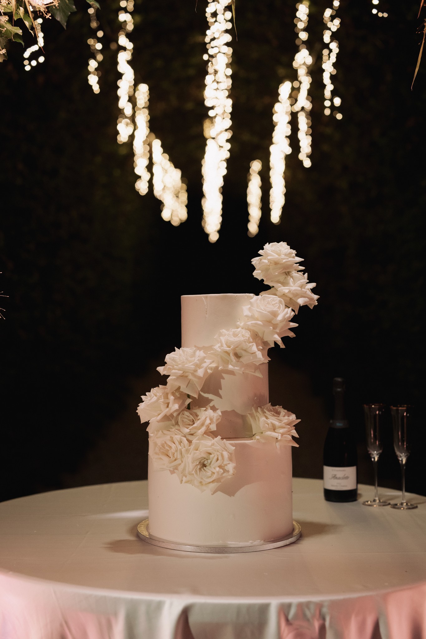 Torta nuziale a tre piani con fiori bianchi sotto lampadari luminosi, ricevimento matrimoniale a Villa Negri