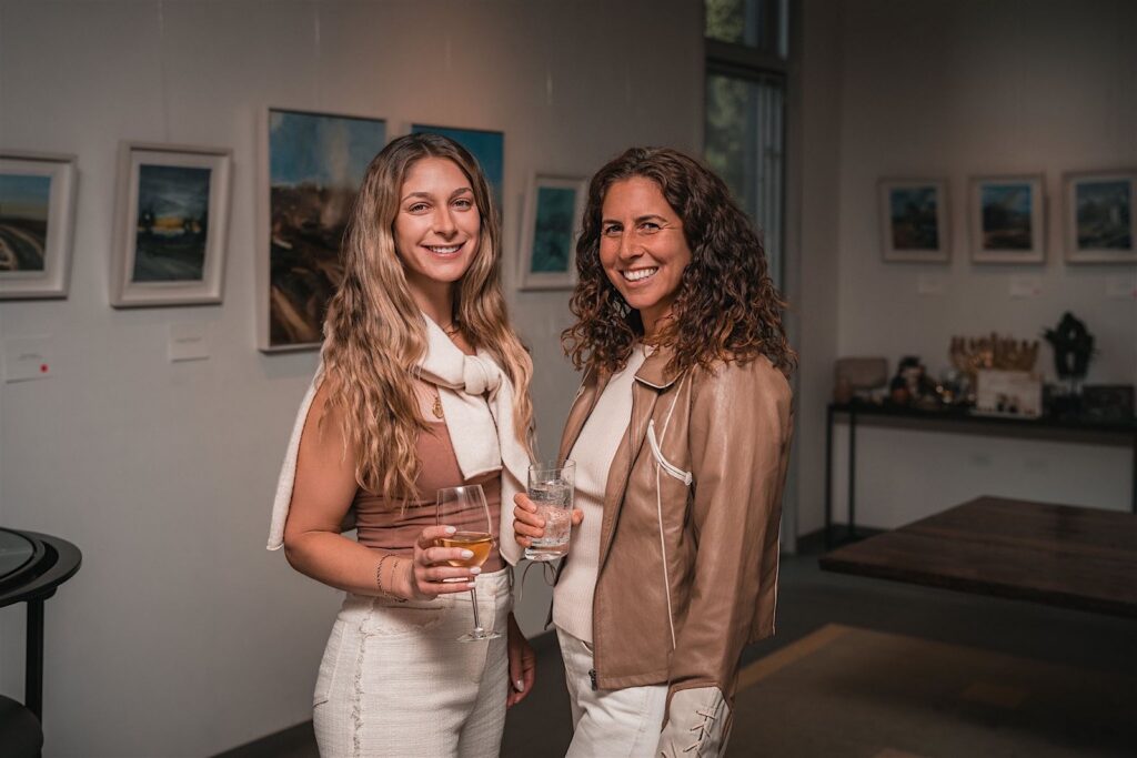 Two smiling guests holding drinks in an art gallery with framed landscape paintings