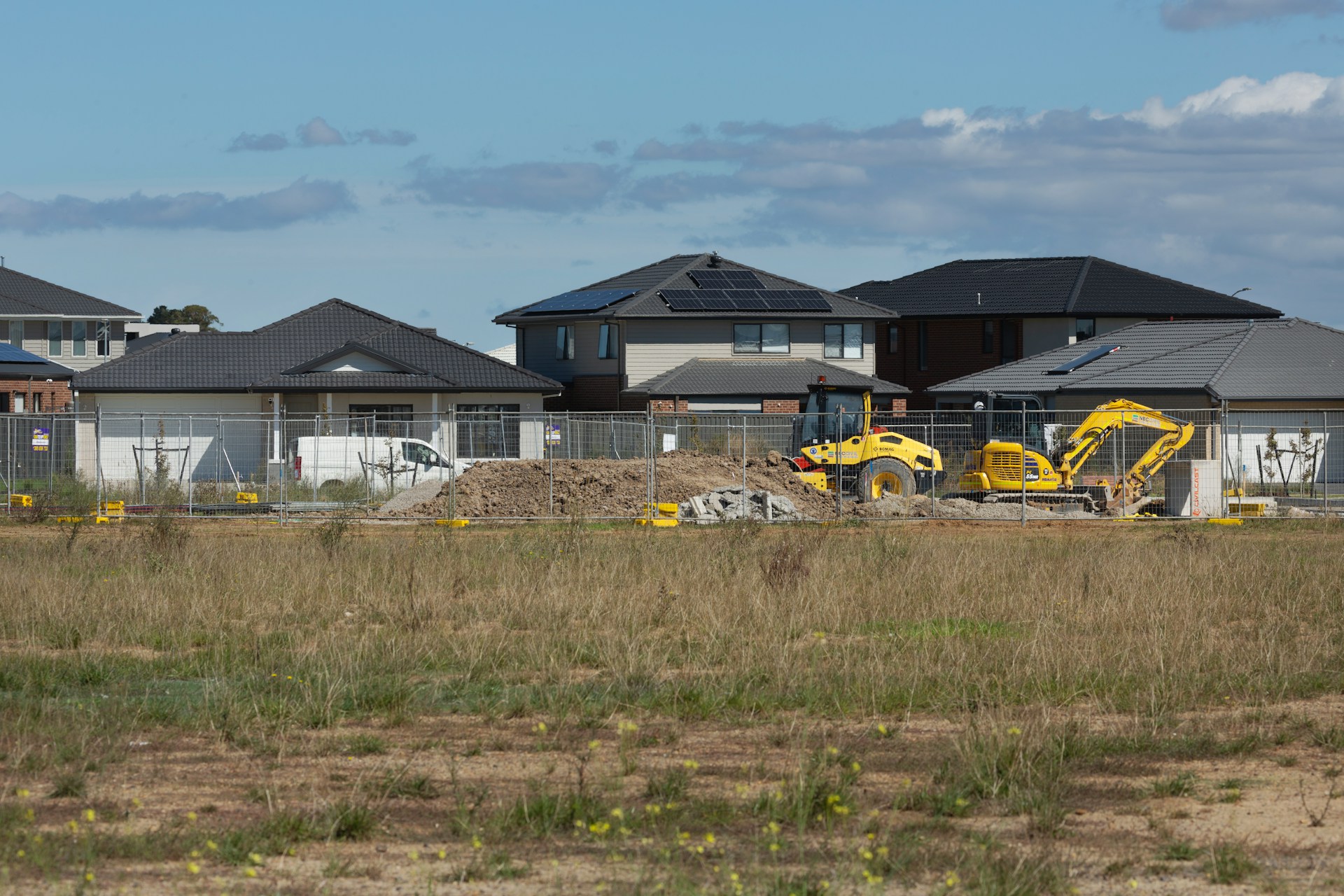 A construction worker in a yellow hard hat and safety goggles uses a drill on wood outside a wooden house under a clear blue sky.