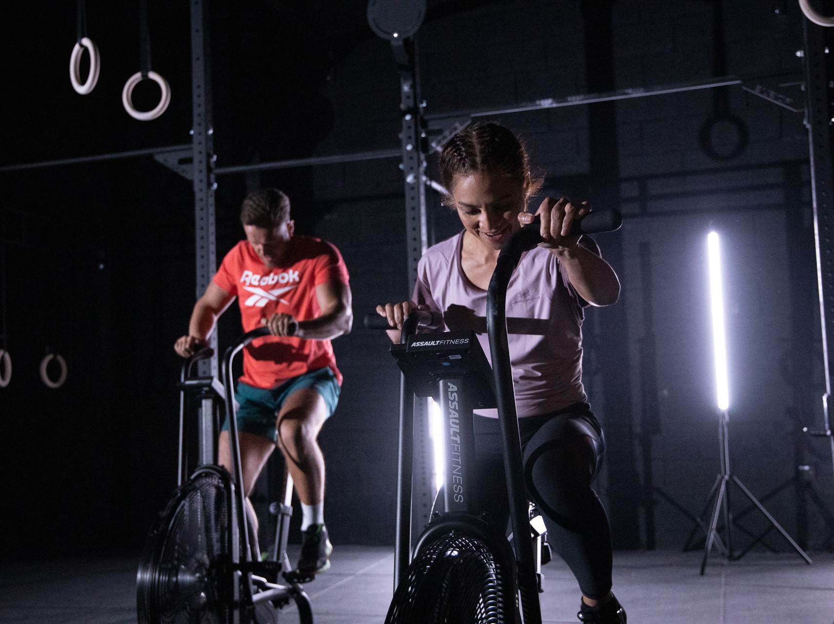 Two athletes training hard on air bikes in a dark gym, lit by a single vertical strip light.
