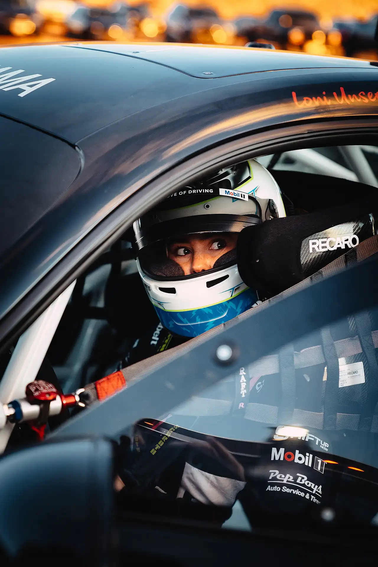 Loni Unser sitting in the driver’s seat of her red Acura race car, looking out the window, with Mobil 1 , Recaro, and Pep Boys logos visible.