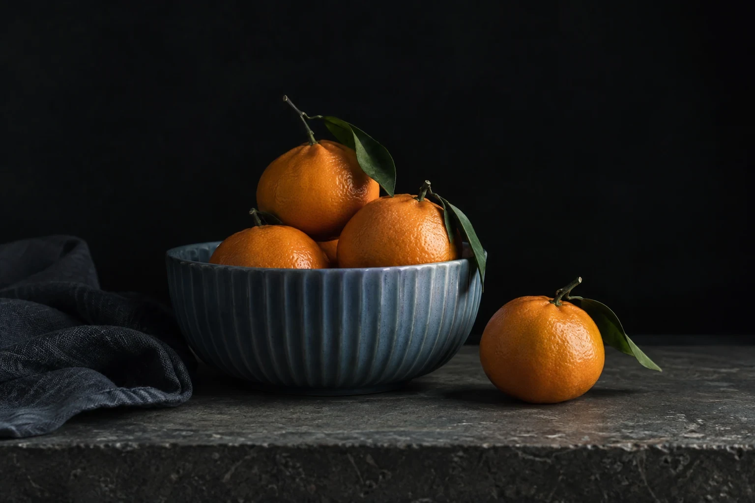Ribbed muted blue bowl holding four tangerines, one fruit resting beside it, near-black background