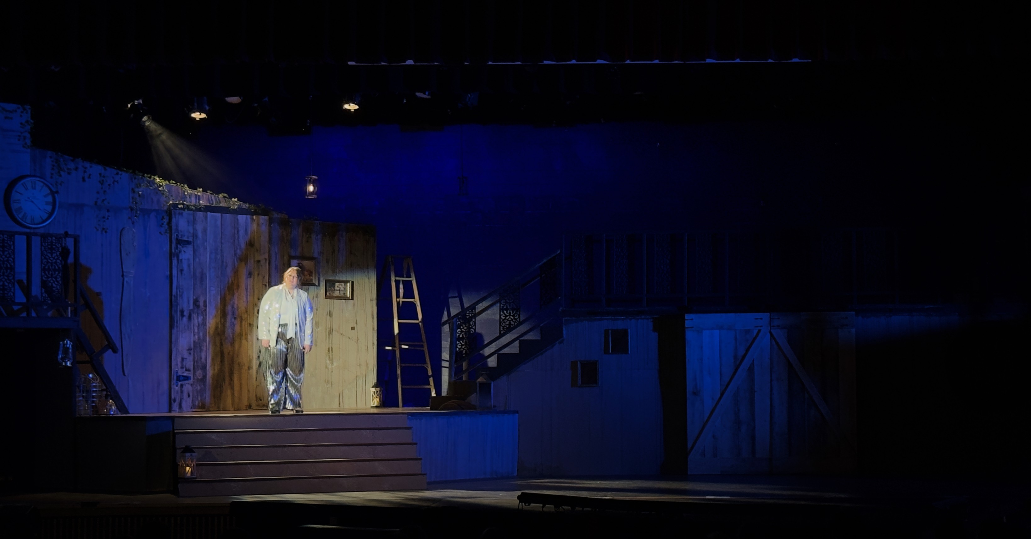 A stage set features a dimly lit scene with minimal stage lighting, spotlighting an individual on a stage right platform against a backdrop of blue-black hues and rustic wooden elements, creating an atmosphere of isolation and focus, enhanced by a breakup gobo effect.