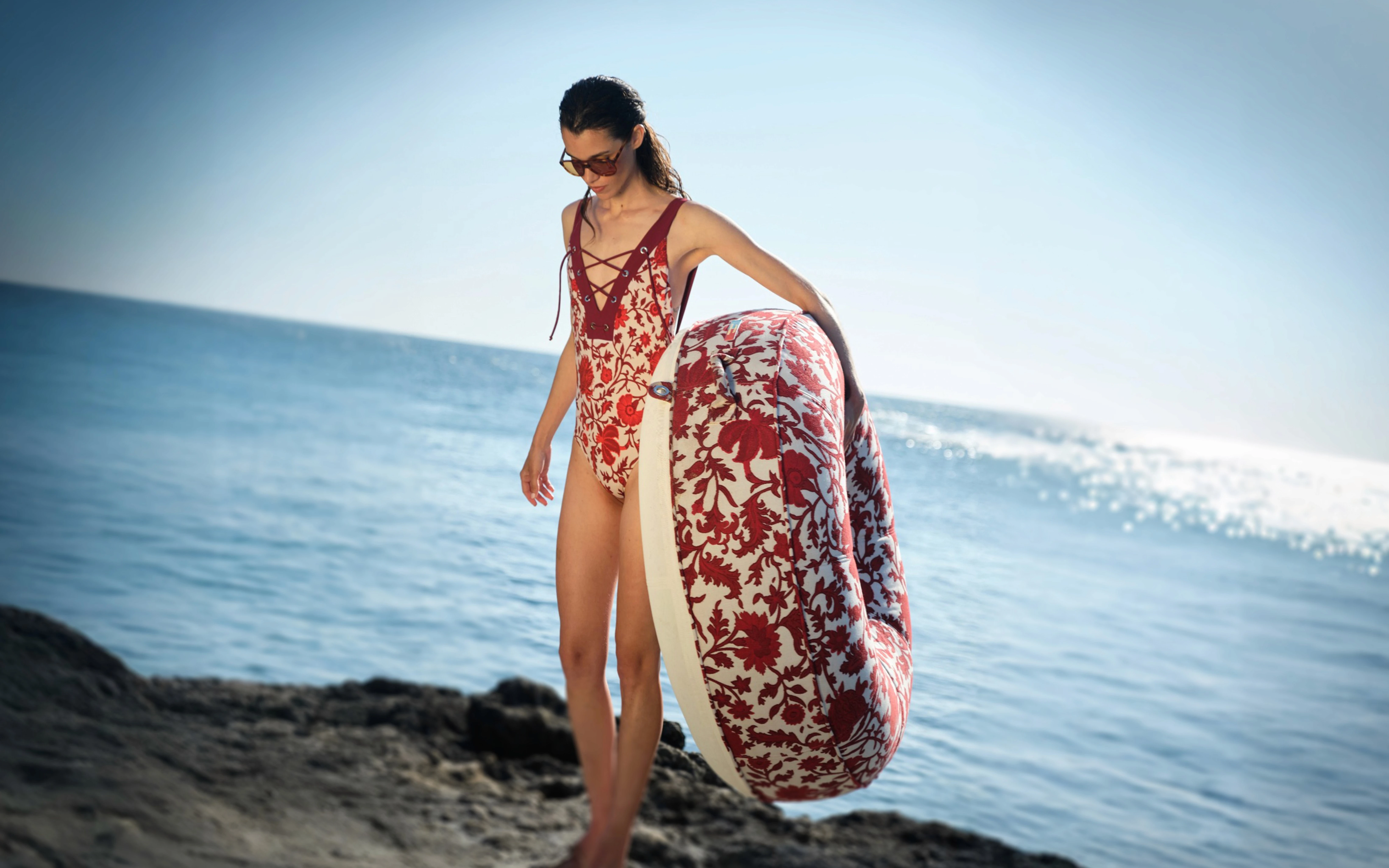 Woman in a matching red floral swimsuit carrying a round luxury pool float with red and cream damask upholstery by Oliver James Lilos — high-end beachside elegance for adults.