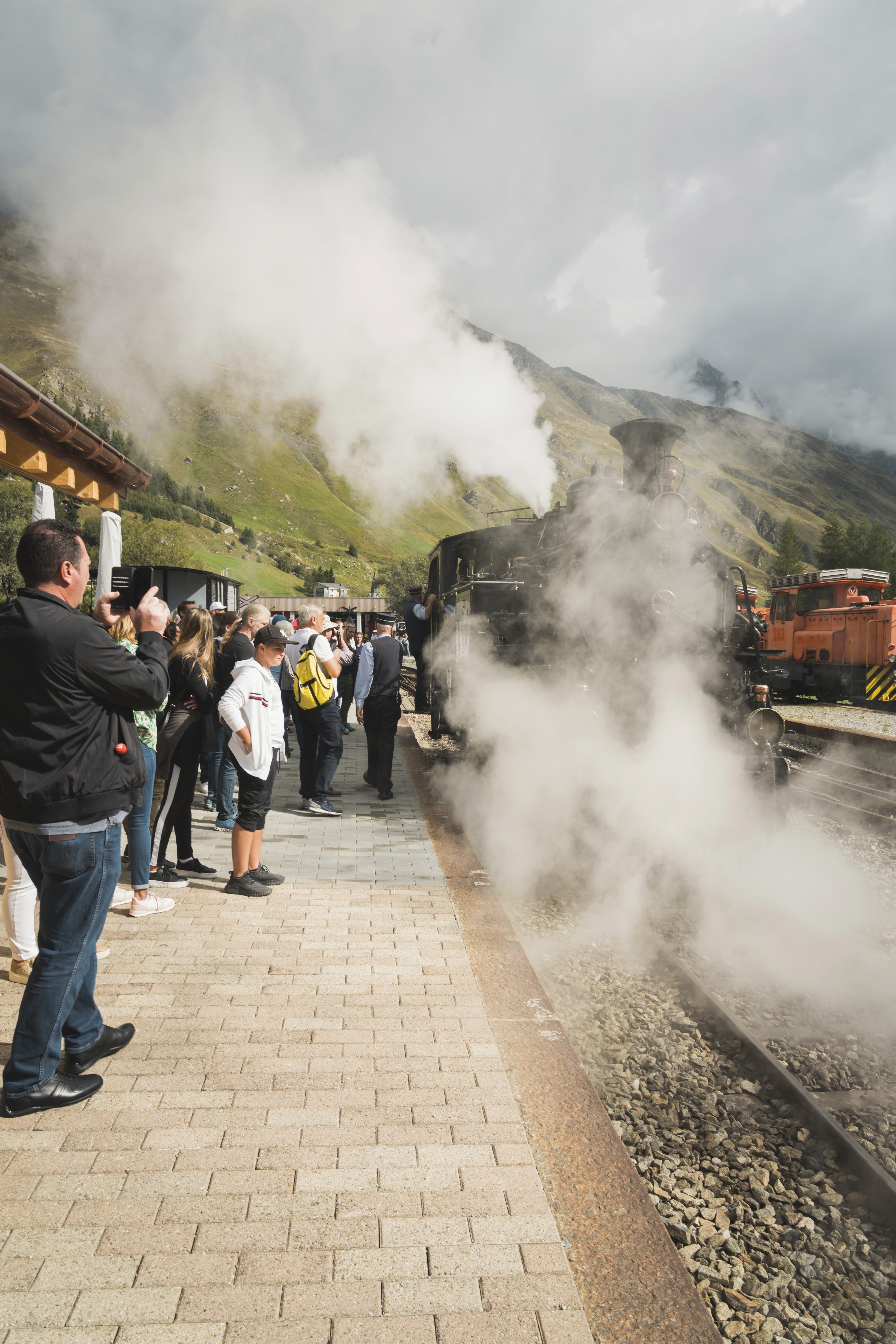 Das Foto zeigt einen gepflasterten Bahnsteig an einem bewölkten Tag, auf dem eine Menschenmenge die Ankunft einer historischen, schwarzen Dampflokomotive beobachtet. Die Lokomotive stößt eine große, dichte Wolke aus weißem Dampf aus, die den Gleisbereich teilweise verhüllt. Im Vordergrund links steht ein Mann in dunkler Jacke und Jeans, der die Szene mit seinem Smartphone filmt oder fotografiert. Unter den Wartenden am Rand des Bahnsteigs befindet sich auch Bahnpersonal in Uniform. Im Hintergrund erheben sich grüne Berghänge unter einem wolkigen Himmel, und auf einem Nebengleis auf der rechten Seite ist eine weitere, orangefarbene Lokomotive zu erkennen.