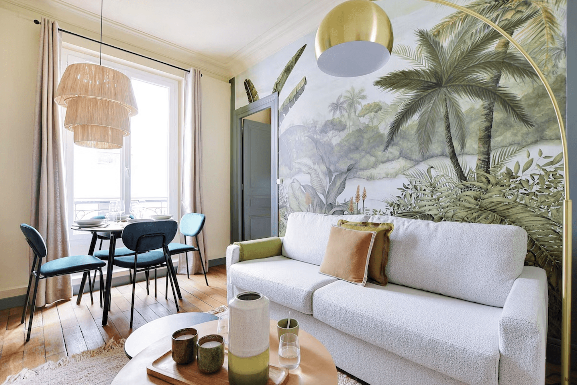 A modern, glass-walled house with warm interior lighting is perched on a rocky cliff overlooking a calm sea at sunset, under a clear blue sky.