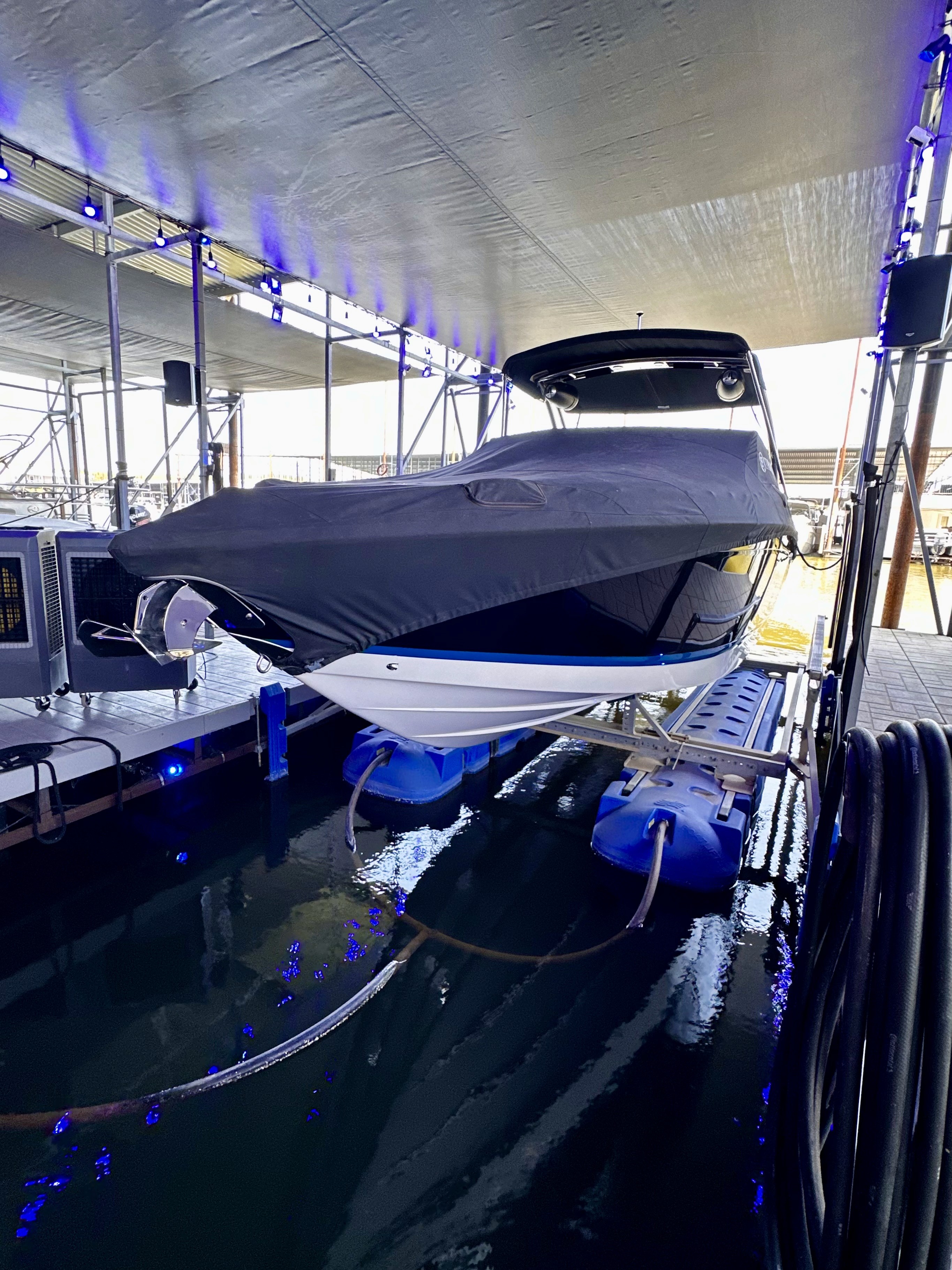A sleek, covered boat is securely docked in a modern marina under a large, protective canopy, with soft blue lighting reflecting off the water, creating a serene and contemporary atmosphere.