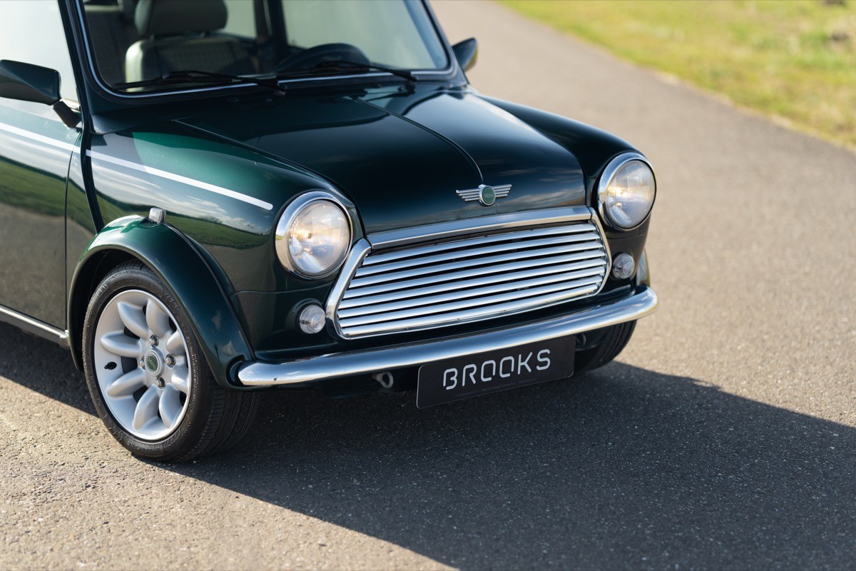 Close-up of a vintage green car’s front end, showcasing its round headlights and chrome grille against a grassy backdrop.